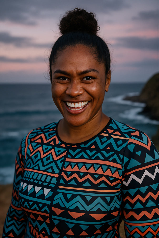 A female surfer stands at the edge of a rocky shoreline under a dusky sky streaked with pastel pink and lavender clouds, wearing a bold tribal-inspired teal and coral long-sleeve rash guard. She designed the “Universe” sticker for Aloha Tinyboards (finger sized mini foam surfboard) as part of their Stories from the Lineup series, capturing the galaxies, space, and planetaries themes.
