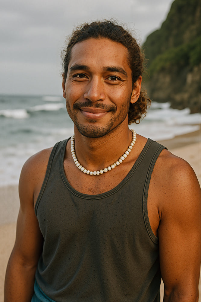 A male surfer-musician stands on a palm-fringed beach at golden hour, wearing a charcoal tank top and chunky puka-shell necklace, his curly hair tied back loosely. He designed the “Music” sticker for Aloha Tinyboards (finger sized mini foam surfboard) as part of their Stories from the Lineup series, blending surf culture with soulful melody.