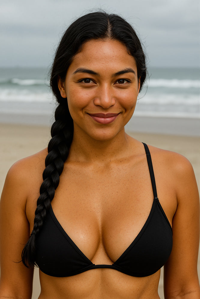 A female surfer stands at the water’s edge under overcast skies in a simple black triangle bikini top, her long dark braid draped over one shoulder. She designed the “Hawaiian Flowers” sticker for Aloha Tinyboards (finger sized mini foam surfboard) as part of their Stories from the Lineup series, evoking serene island elegance.