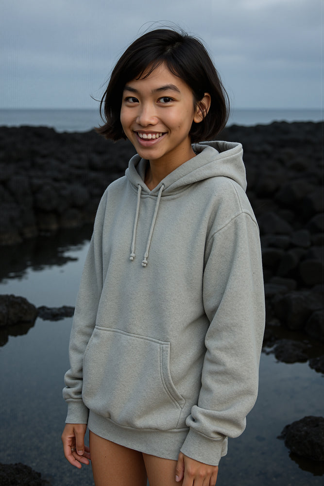 A teenage surfer poses on black lava rocks by the sea, sticking out her tongue and crossing her eyes mid-laugh, wearing a dusty rose T-shirt and beige drawstring shorts. She designed the “Tiger” sticker for Aloha Tinyboards (finger sized mini foam surfboard) as part of their Stories from the Lineup series, infusing playful energy into the lineup.