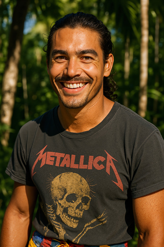 A male surfer stands in a sun-dappled palm grove at midday, smiling broadly in a faded black Metallica T-shirt and colorful geometric-print board shorts. He designed the “Geometric” sticker for Aloha Tinyboards (finger sized mini foam surfboard) as part of their Stories from the Lineup series, showcasing bold pattern and island style.