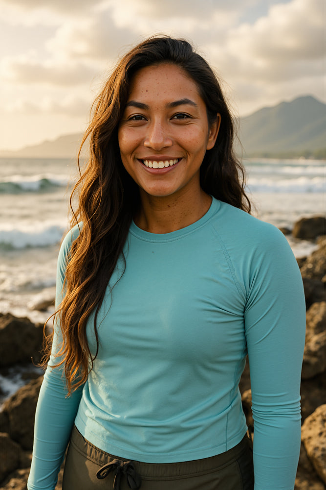 A female surfer gazes across rocky tide pools at golden hour, wearing a teal long-sleeve rash guard and dark board shorts, her loose waves catching the sunset light. She designed the “Weave” sticker for Aloha Tinyboards (finger sized mini foam surfboard) as part of their Stories from the Lineup series, infusing woven pattern motifs and warm island hues into the collection.