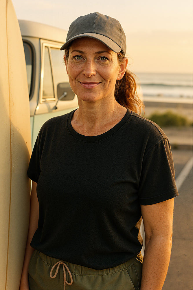 A female surfer leans against a pastel-striped surfboard on a sandy beach at sunset, wearing a charcoal cap, black tee, and olive-green shorts as the ocean breeze lifts her ponytail. She designed the “Boho Plants” sticker for Aloha Tinyboards (finger sized mini foam surfboard) as part of their Stories from the Lineup series, blending bohemian botanical elements with serene coastal vibes.
