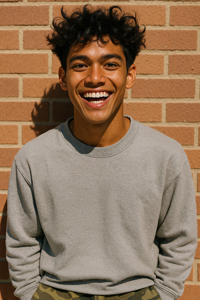A male surfer stands against a red brick wall under bright midday sun, wearing a light gray sweatshirt and camo-print pants, his dark curls tousled by a light breeze. He designed the “Dripping” sticker for Aloha Tinyboards (finger sized mini foam surfboard) as part of their Stories from the Lineup series, adding youthful street-style flair to the collection.